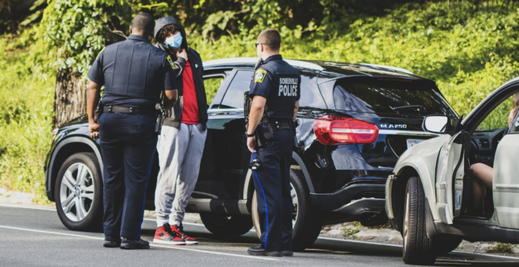 Police officers question a masked individual during a roadside stop beside two cars.